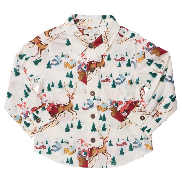 A long-sleeve button-up shirt with a festive Christmas print featuring Santa Claus in red sleighs, brown reindeer, green pine trees, and small houses on an off-white background.