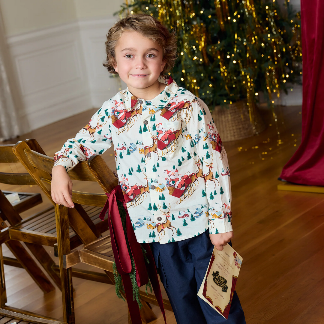 A young boy wearing a festive holiday-themed long-sleeve hooded sweatshirt and navy pants, standing in a decorated room with a Christmas tree and wooden chairs in the background.