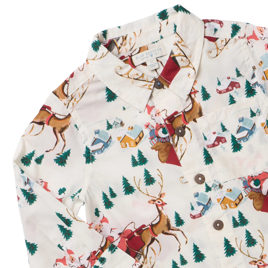 A close-up of a long-sleeve button-up shirt with a festive Christmas village print featuring reindeer, Santa Claus, snow trees, and houses on an off-white background.