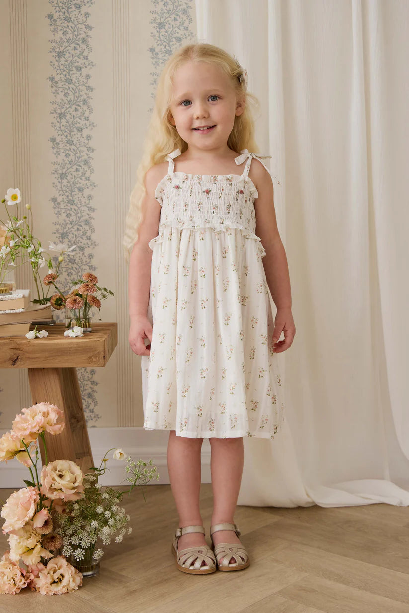 Young girl in a floral dress standing in a room with flowers and a wooden table.