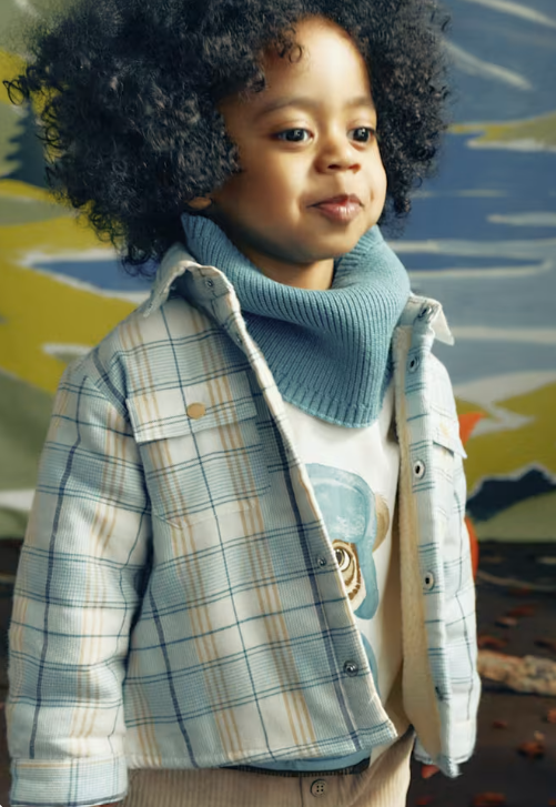 A toddler with curly black hair wearing a light blue and white plaid overshirt over a white graphic t-shirt and a light blue knit cowl neck warmer, posed against a colorful painted background.