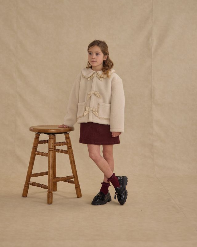 Young girl in a beige coat and maroon skirt standing next to a wooden stool on a beige background
