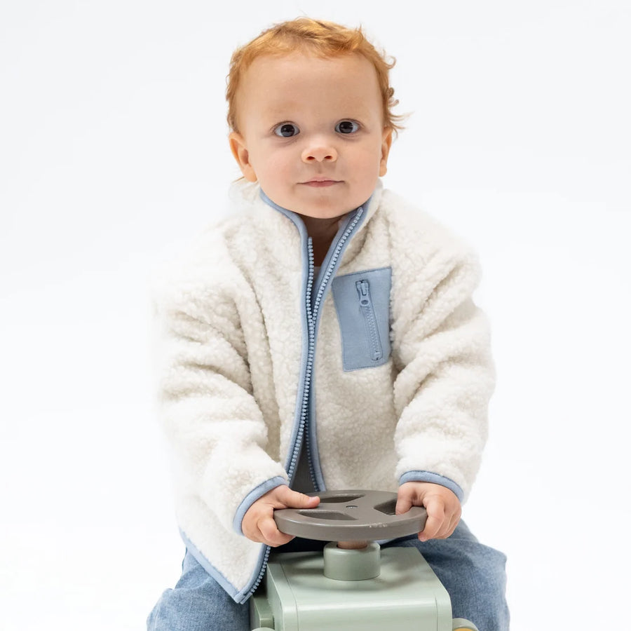 A baby with red hair wearing a cream sherpa fleece zip-up jacket with light blue trim and a matching blue chest pocket, sitting on a mint green toy vehicle against a white background.