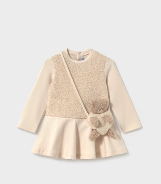 A toddler girl's long-sleeve dress in cream and beige, featuring a textured sherpa-style front panel, a flared skirt, and a small plush teddy bear accessory hanging from the shoulder strap, shown on a light gray background.