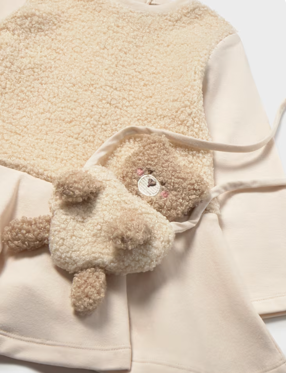 A close-up of a cream-colored fleece garment with a small beige teddy bear plush accessory featuring embroidered facial details and a drawstring strap.