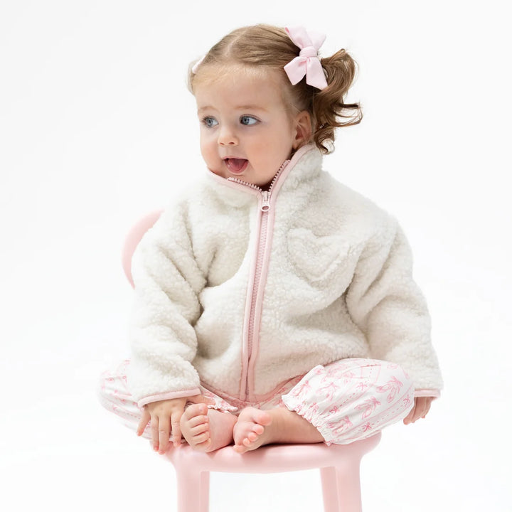A toddler girl sitting on a small pink chair, wearing a cream-colored sherpa jacket with a pink zipper and matching pink patterned pants, with a pink bow in her hair.