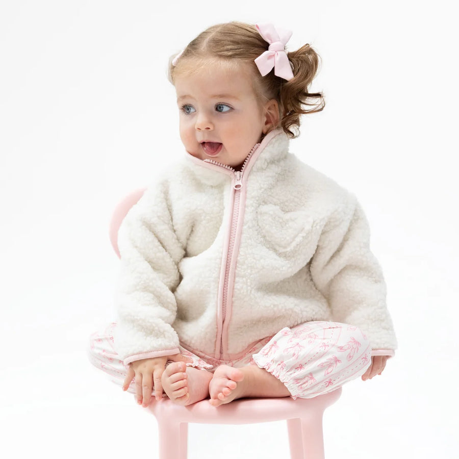 A toddler girl sitting on a small pink chair, wearing a cream-colored sherpa jacket with a pink zipper and matching pink patterned pants, with a pink bow in her hair.