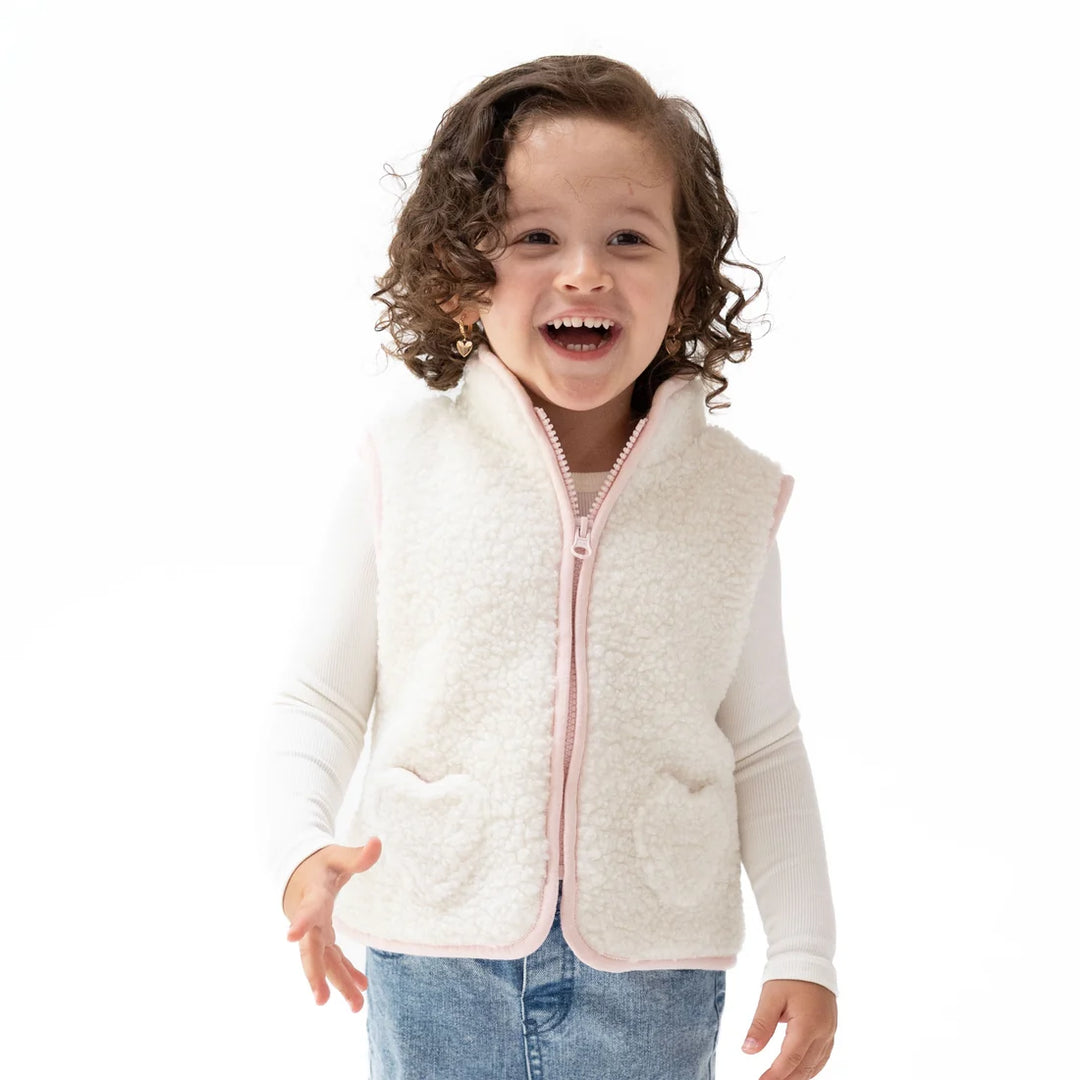 A young girl with curly brown hair smiling and laughing while wearing a cream sherpa zip-up vest with light pink trim over a white long-sleeve shirt and blue jeans, set against a plain white background.