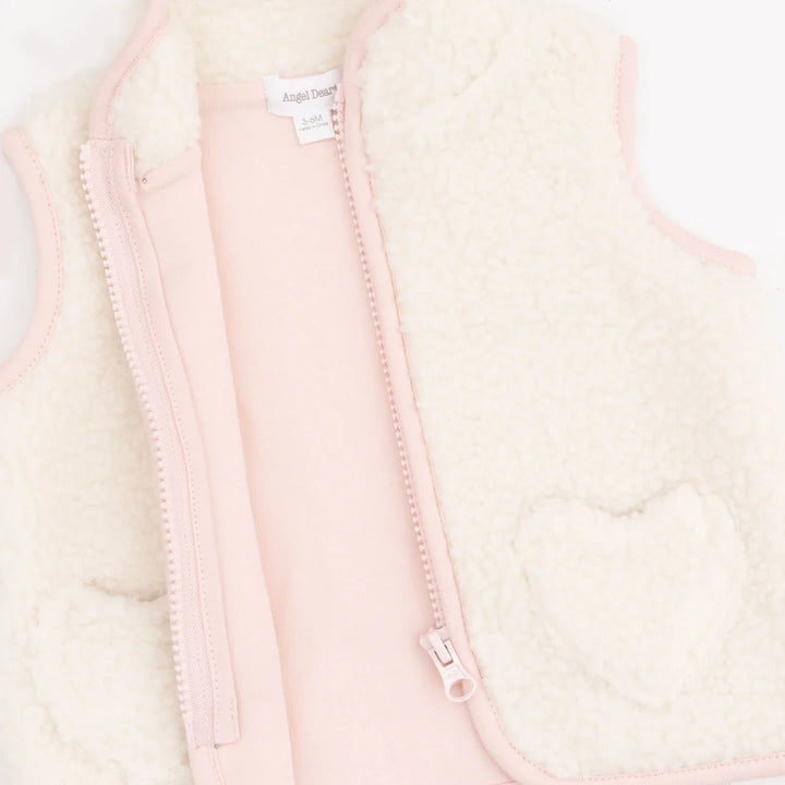 A close-up of a children's zip-front vest with a soft cream sherpa lining and pale pink outer fabric, zipper, and trim. A heart-shaped patch is visible on the right side, and a small interior label reads 'Angel Dear'.