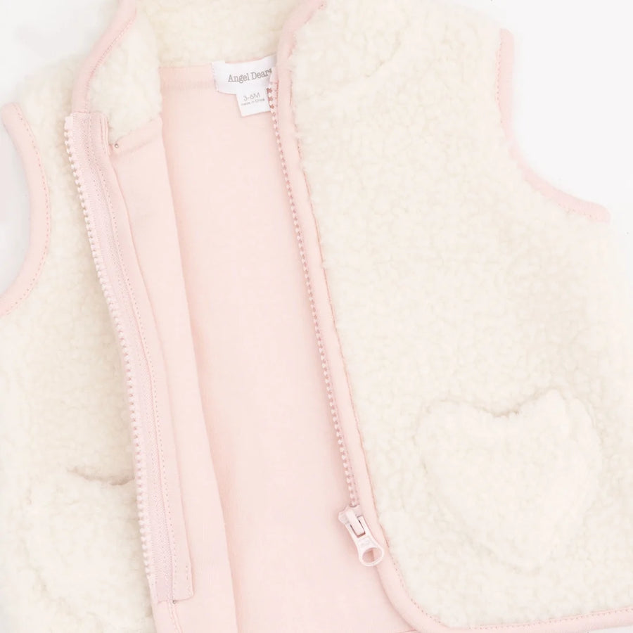 A close-up of a children's zip-front vest with a soft cream sherpa lining and pale pink outer fabric, zipper, and trim. A heart-shaped patch is visible on the right side, and a small interior label reads 'Angel Dear'.