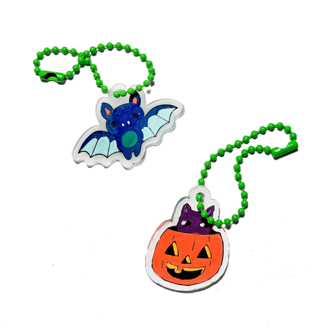 Two Halloween-themed acrylic keychains on green beaded chains, featuring a blue bat and an orange jack-o'-lantern pumpkin with a purple witch hat, set against a plain white background.