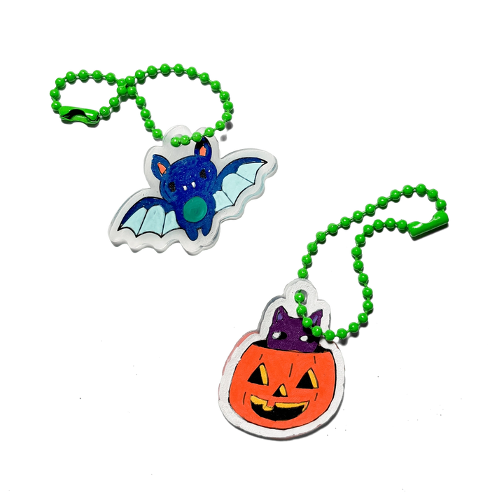 Two Halloween-themed acrylic keychains on green beaded chains, featuring a blue bat and an orange jack-o'-lantern pumpkin with a purple witch hat, set against a plain white background.