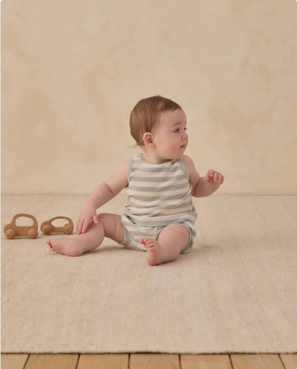 Baby sitting on a beige carpet wearing a striped romper with wooden toys in the background.