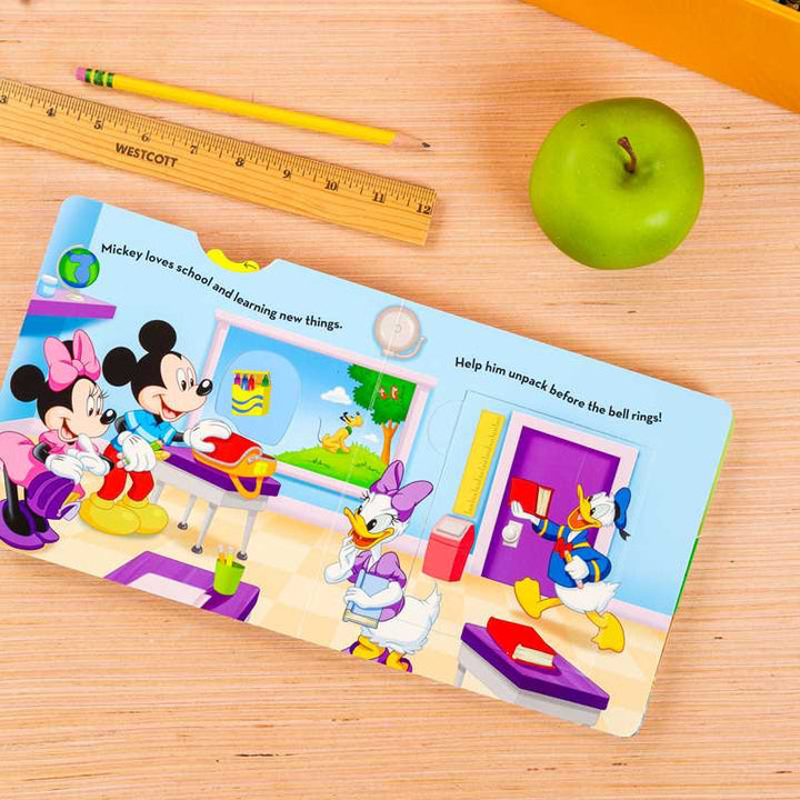 An open children's picture book featuring Mickey Mouse, Minnie Mouse, Daisy Duck, and Donald Duck in a colorful school scene, placed on a light wood tabletop with a yellow ruler, pencil, and green apple nearby.