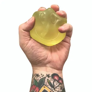 A hand with a floral tattoo is holding a translucent yellow-green, smiley-face-shaped stress toy against a plain white background.