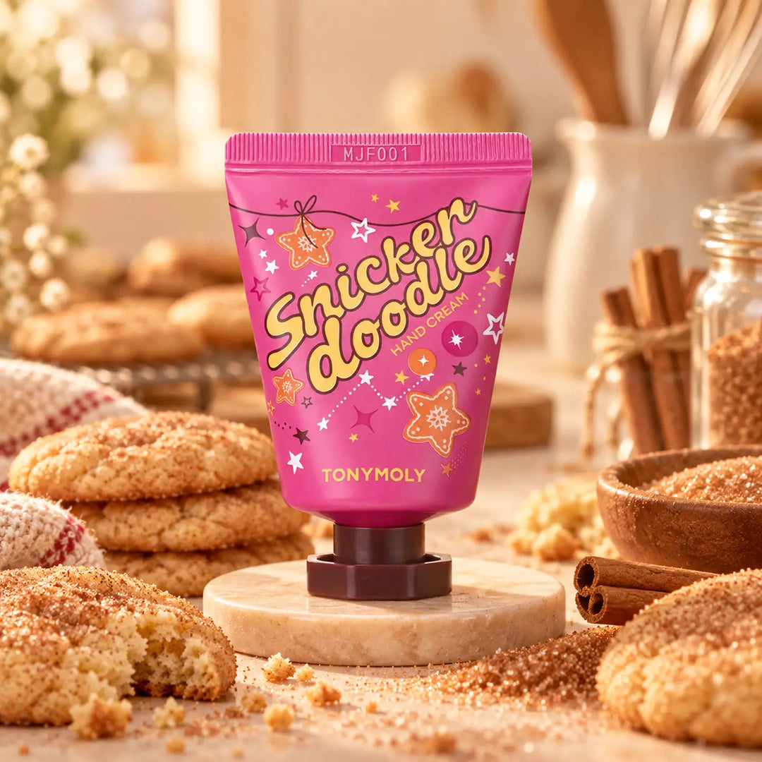 A pink squeeze tube of TONYMOLY Snickerdoodle hand cream, styled on a wooden stand surrounded by sugar cookies, cinnamon sticks, and warm spices in a cozy, warm-toned setting.
