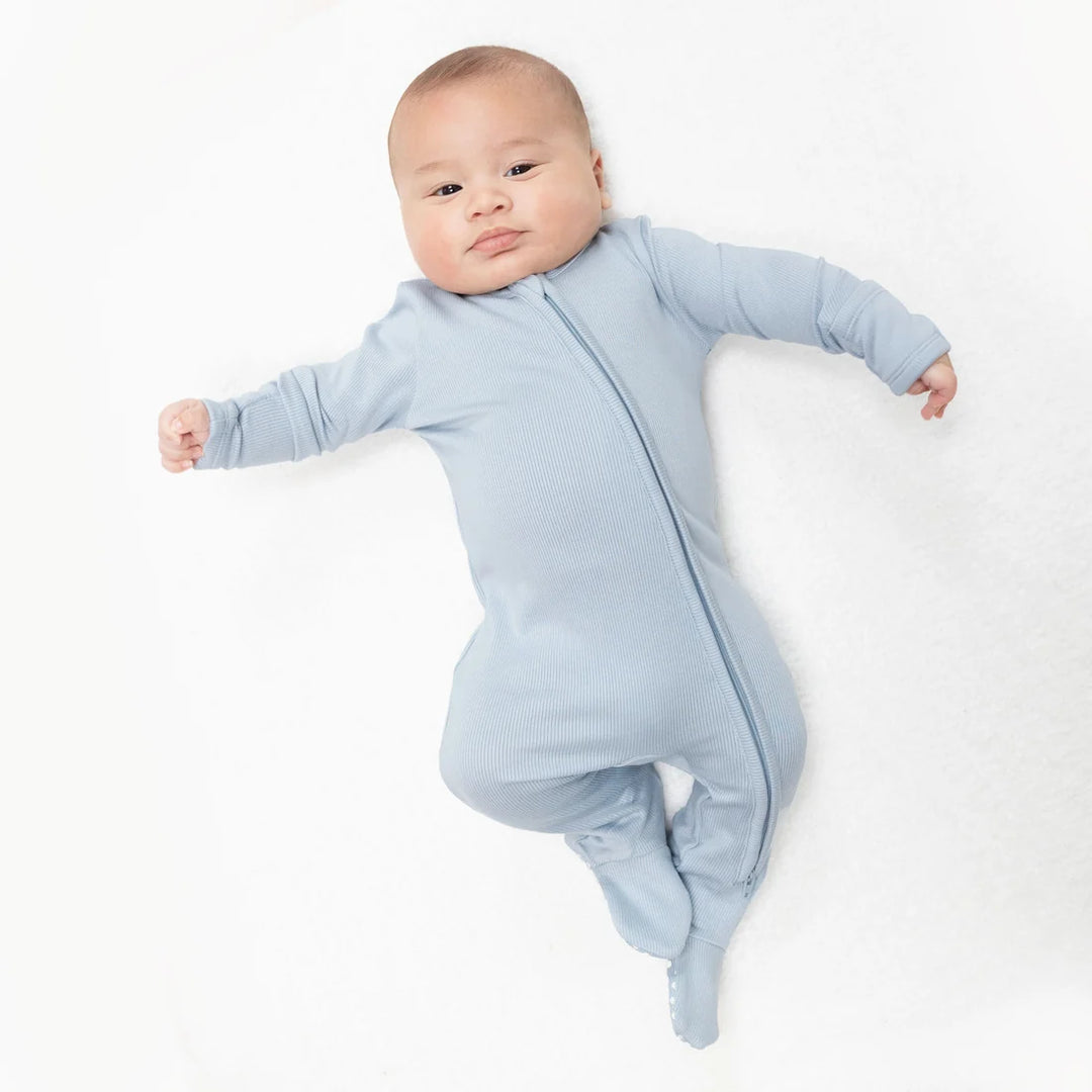 A baby lying on a white textured blanket, wearing a light blue long-sleeve footed sleeper with a front zipper.