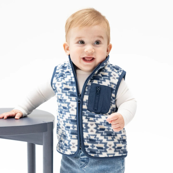 A baby standing behind a gray table, wearing a navy and white geometric patterned zip-up vest over a white long-sleeve shirt and blue jeans.
