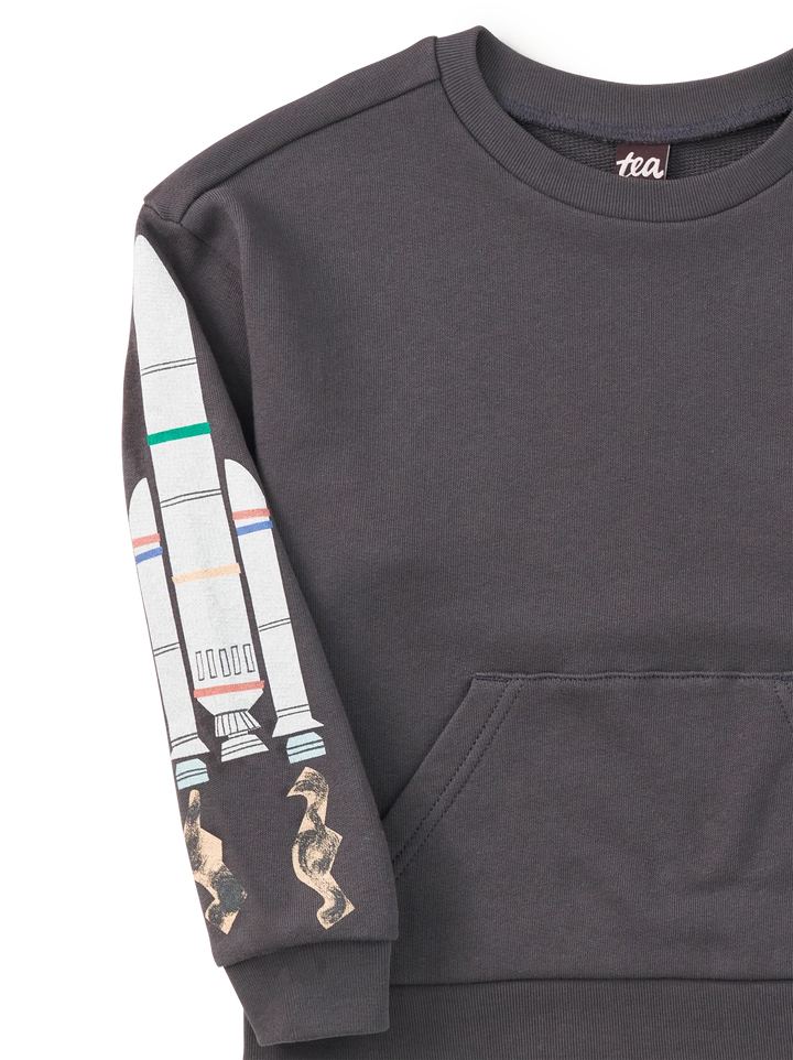 A close-up of a dark gray crewneck sweatshirt featuring a graphic print of a rocket ship on one sleeve, with two small seahorse figures near the cuff. The brand name 'tea' is visible on the inner neck label.