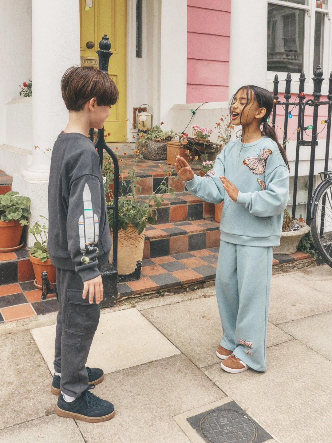 Two children standing on a front porch wearing matching light blue and dark gray sweatshirt and sweatpants sets, with a house, potted plants, and a bicycle in the background.