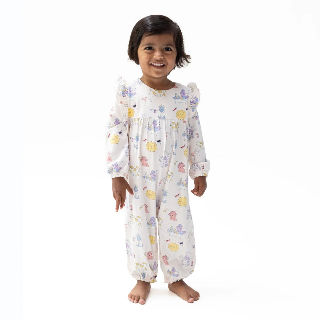 A smiling toddler girl stands barefoot against a plain white studio background, wearing a long-sleeve smocked pajama romper with a white base and a scattered pastel spa dogs print.