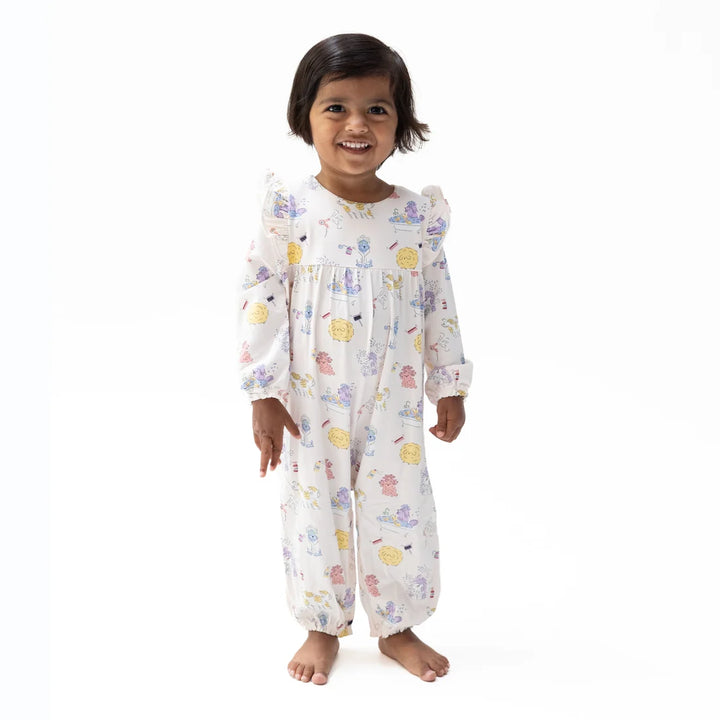 A smiling toddler girl stands barefoot against a plain white studio background, wearing a long-sleeve smocked pajama romper with a white base and a scattered pastel spa dogs print.