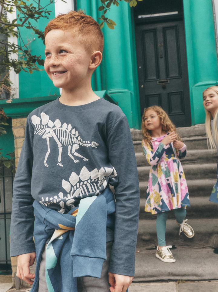 A young boy with red hair smiling and looking to the side, wearing a slate blue long-sleeve shirt with a white dinosaur skeleton graphic. He has a blue jacket tied around his waist, standing on stone steps in front of a bright green building with other children in the background.