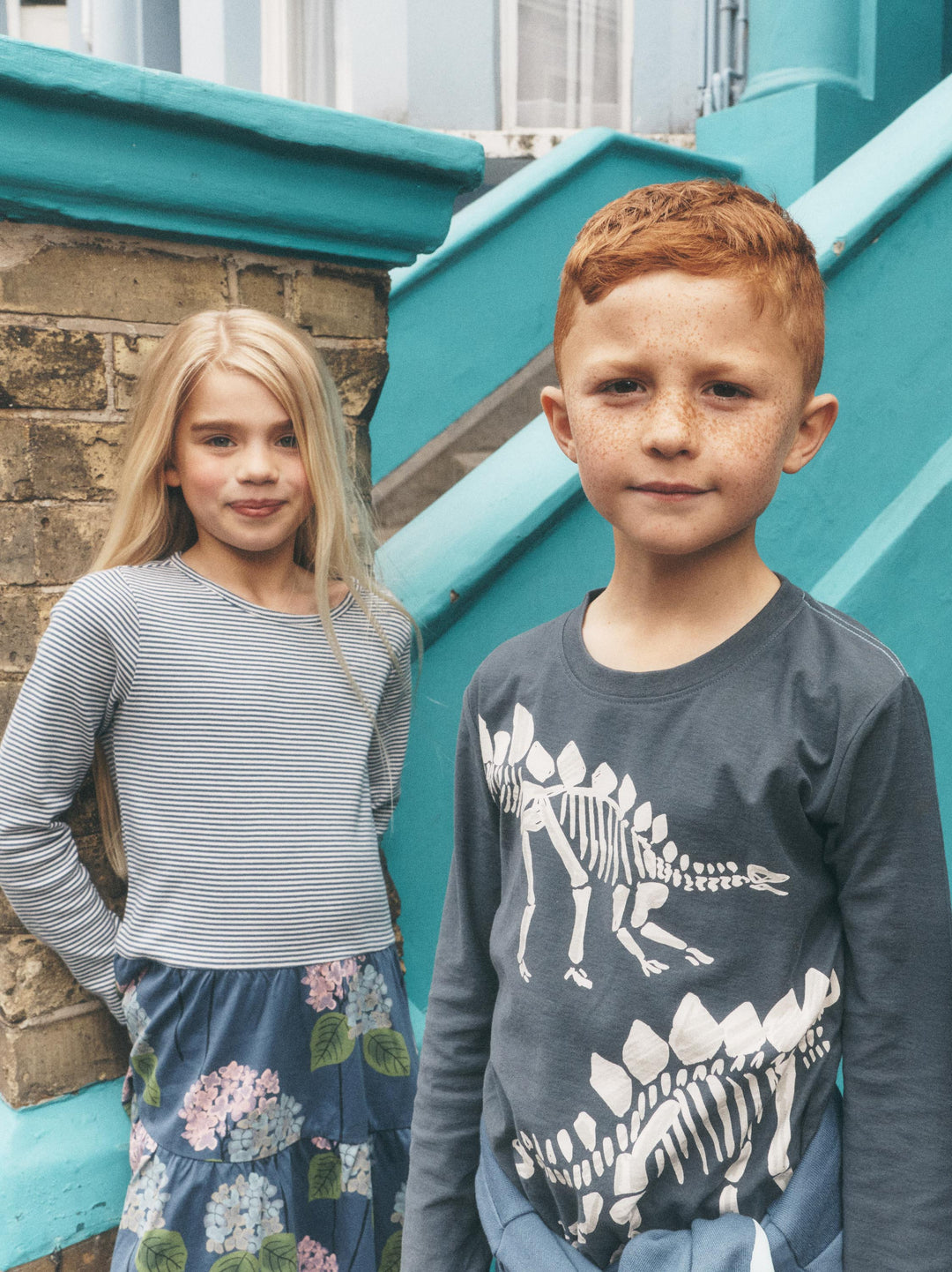 A young boy with red hair and a girl with long blonde hair standing outdoors in front of a turquoise staircase. The boy is wearing a dark gray long-sleeve shirt with white dinosaur skeleton prints, and the girl is wearing a gray and white striped long-sleeve top with a blue floral skirt.