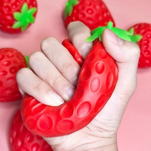 A hand squeezing a red strawberry-shaped squishy toy with green leafy tops against a pink background, with additional strawberry-shaped objects blurred around it.