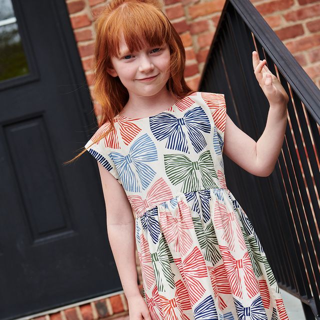 Young girl wearing a colorful dress with bow pattern standing in front of a black door.