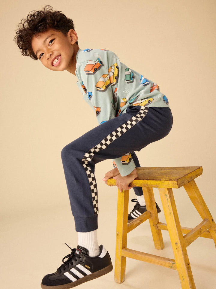 A young boy wearing a light blue long-sleeve shirt with a colorful car print and navy track pants with a black-and-white checkered stripe, sitting on a yellow wooden stool against a beige background.