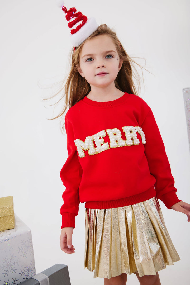 Young girl wearing a red sweater with 'MERRY' and a gold skirt, standing against a white background.