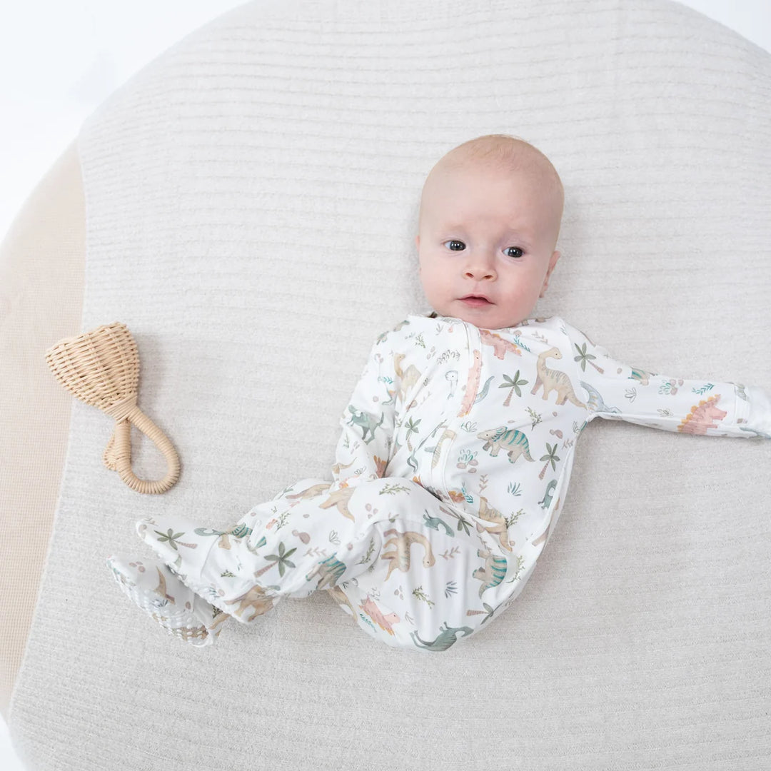 A baby lying on a textured white blanket next to a woven rattan teething toy. The baby is wearing a white footed sleeper with a colorful dinosaur and plant pattern.