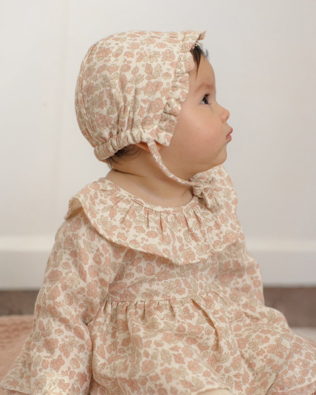 A baby wearing a matching beige and dusty pink floral bonnet and long-sleeve dress, shown in profile against a soft off-white background.