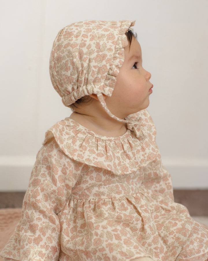A baby wearing a matching beige and dusty pink floral bonnet and long-sleeve dress, shown in profile against a soft off-white background.