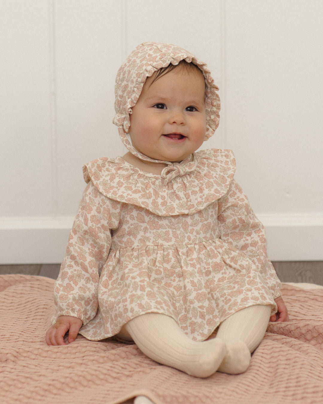 A baby wearing a light beige and pink floral print dress with a large ruffled collar, matching bonnet, and cream tights, sitting on a pink quilted blanket against a white paneled wall.