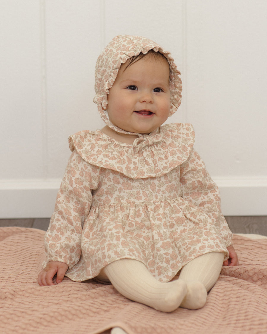 A baby wearing a light beige and pink floral print dress with a large ruffled collar, matching bonnet, and cream tights, sitting on a pink quilted blanket against a white paneled wall.