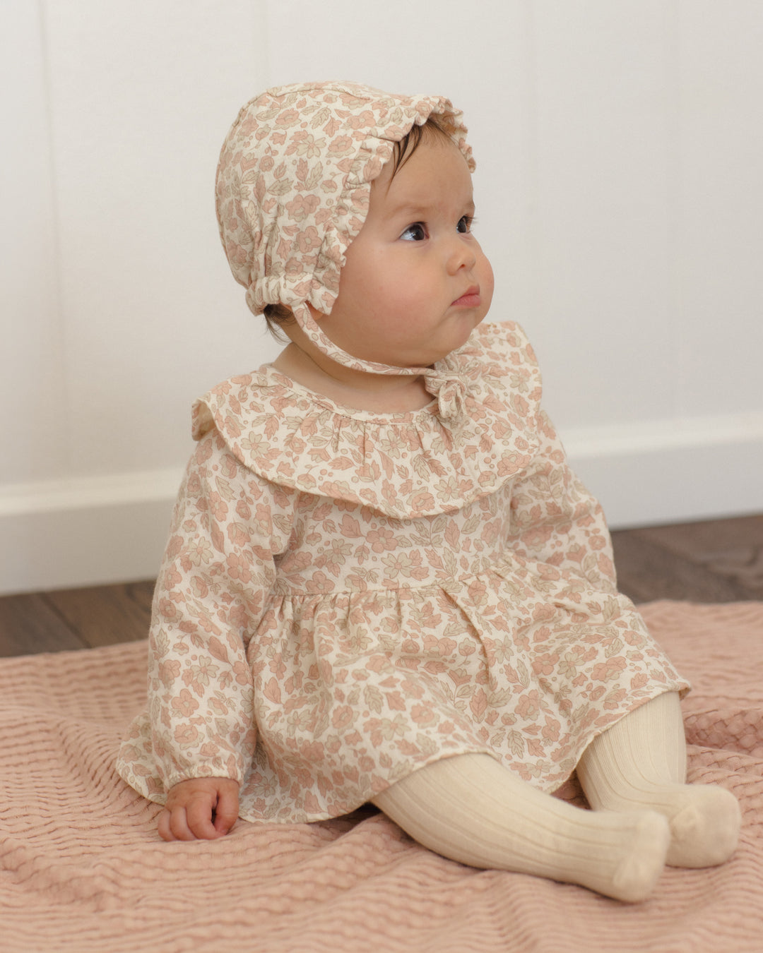 A baby wearing a beige and pink floral print bonnet, matching long-sleeve dress, and cream ribbed tights, sitting on a pink textured blanket.