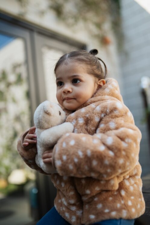 Child in a brown coat with white polka dots holding a stuffed animal outdoors.