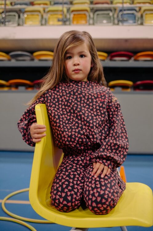 Child wearing a patterned outfit sitting on a yellow chair in an indoor setting with colorful seats in the background.