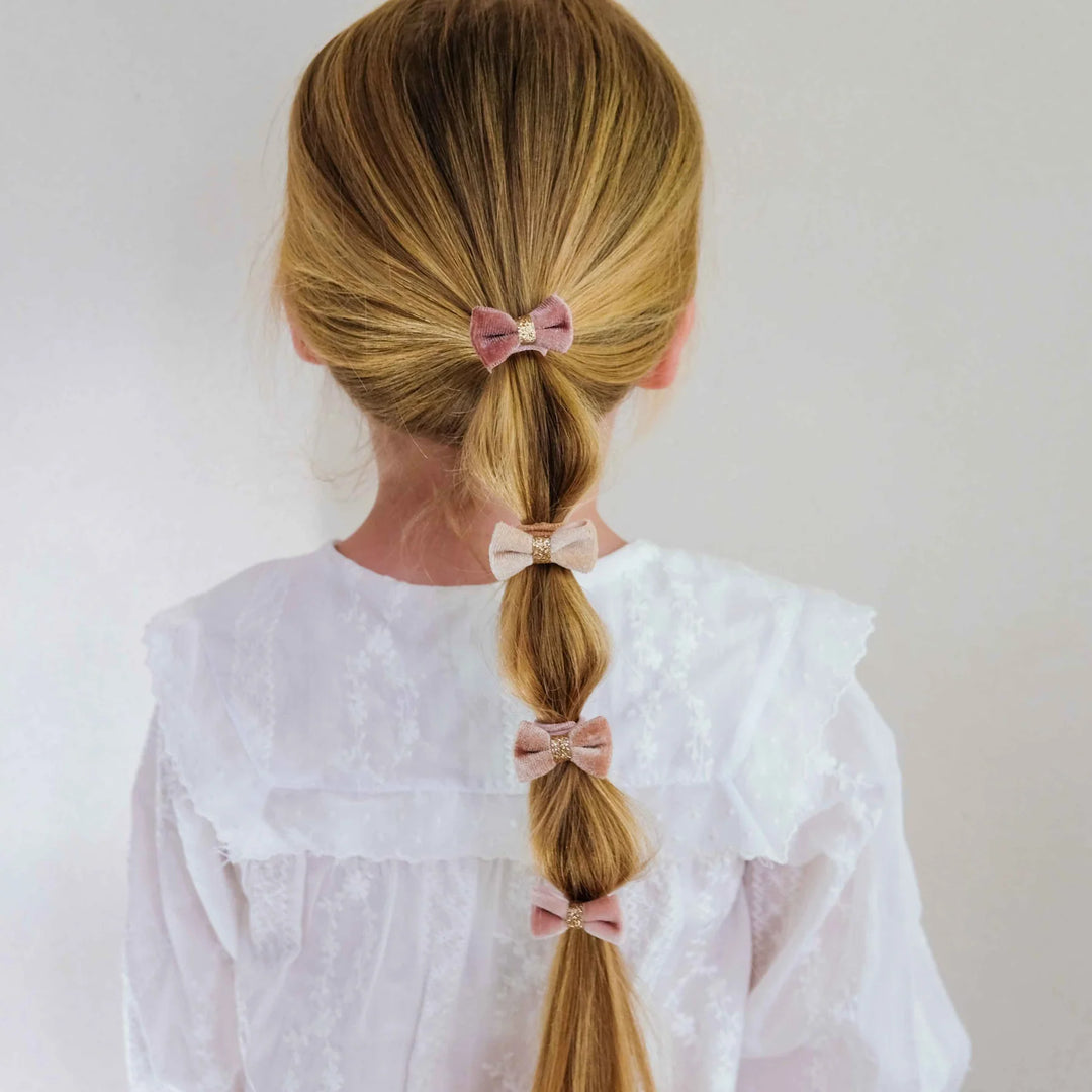 A young girl viewed from behind, wearing a white lace blouse and showcasing a blonde ponytail styled with three pink fabric hair bows.
