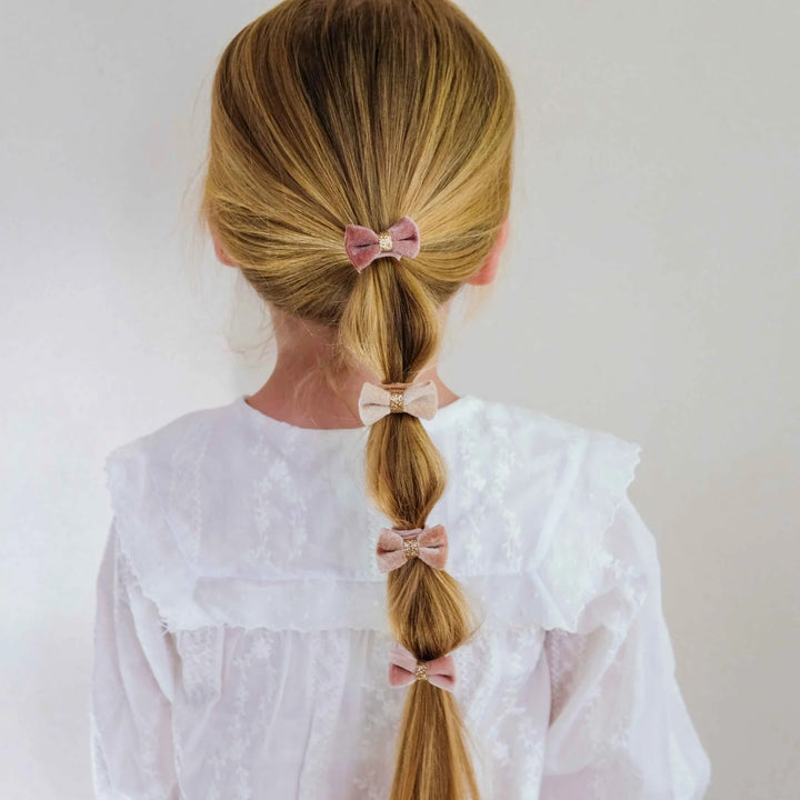 A young girl viewed from behind, wearing a white lace blouse and showcasing a blonde ponytail styled with three pink fabric hair bows.