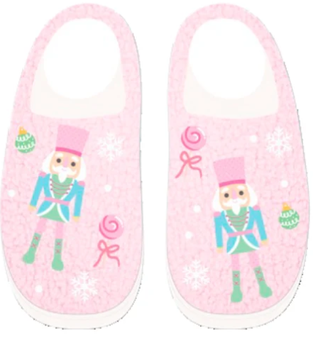 A pair of pink slip-on children's slippers decorated with a nutcracker, snowflakes, candy canes, and ornaments on a light pink background.