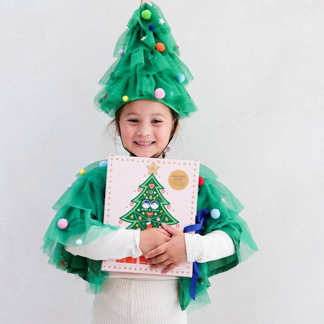Child wearing a green Christmas tree costume holding a book with a Christmas tree design.