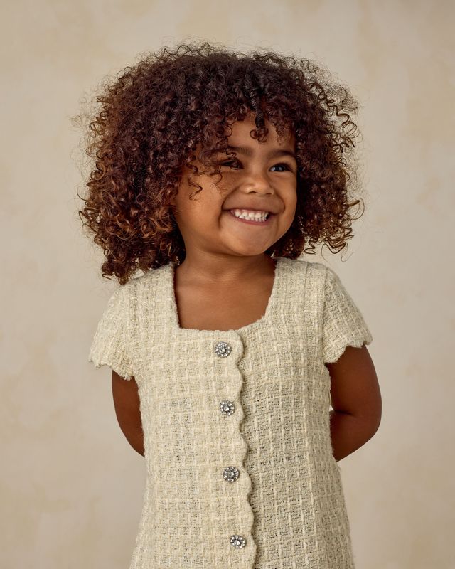 Young girl wearing a light-colored tweed dress with rhinestone buttons against a beige background