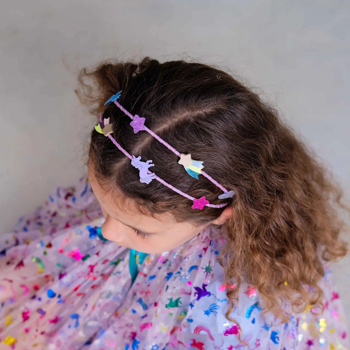 A close-up of a young girl with curly brown hair wearing a pink elastic headband decorated with colorful felt unicorn and star shapes, paired with a sparkly pastel tulle dress.