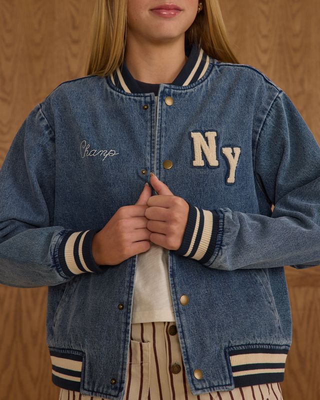 Person wearing a blue denim letterman jacket with 'NY' and 'Champs' text on a wooden background