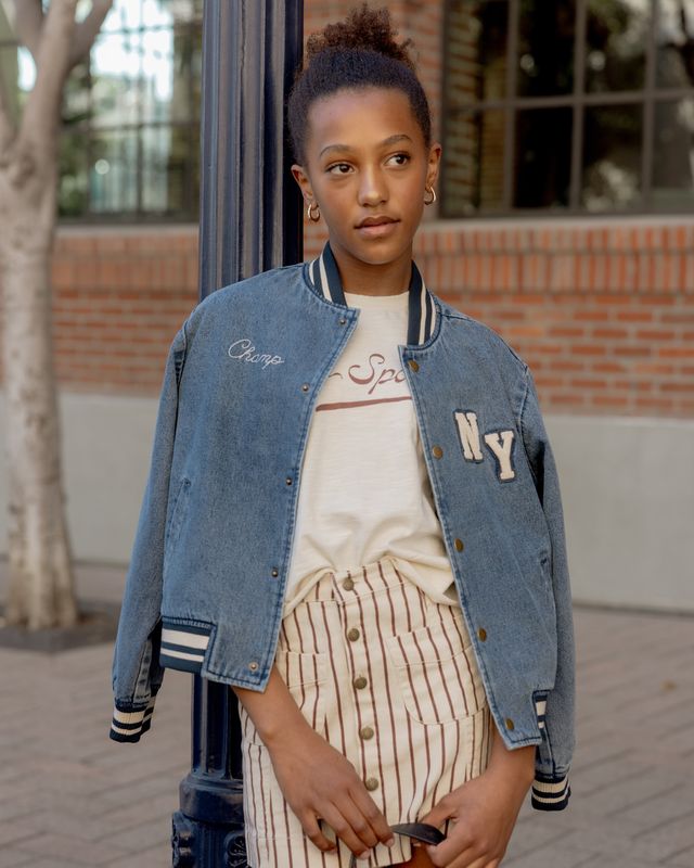 Young girl wearing a denim jacket with 'Champs' and 'NY' patches, standing outdoors.