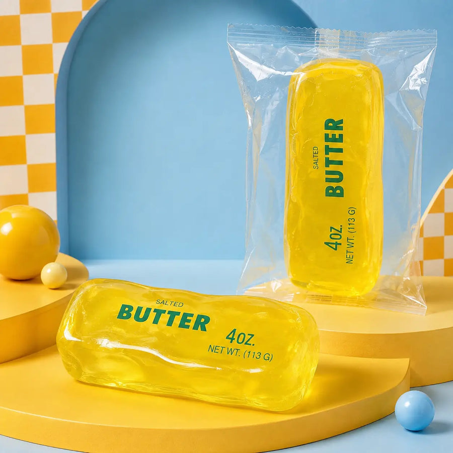Two yellow butter bars in clear plastic packaging, labeled 'SALTED BUTTER 4oz', displayed on yellow geometric platforms against a blue and yellow studio background.