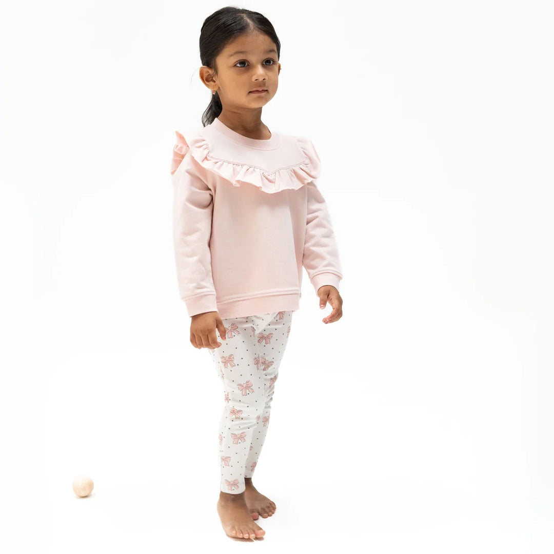 A young girl stands barefoot against a white studio background wearing a light pink long-sleeve sweatshirt with a ruffled collar and white leggings with a pink floral print.